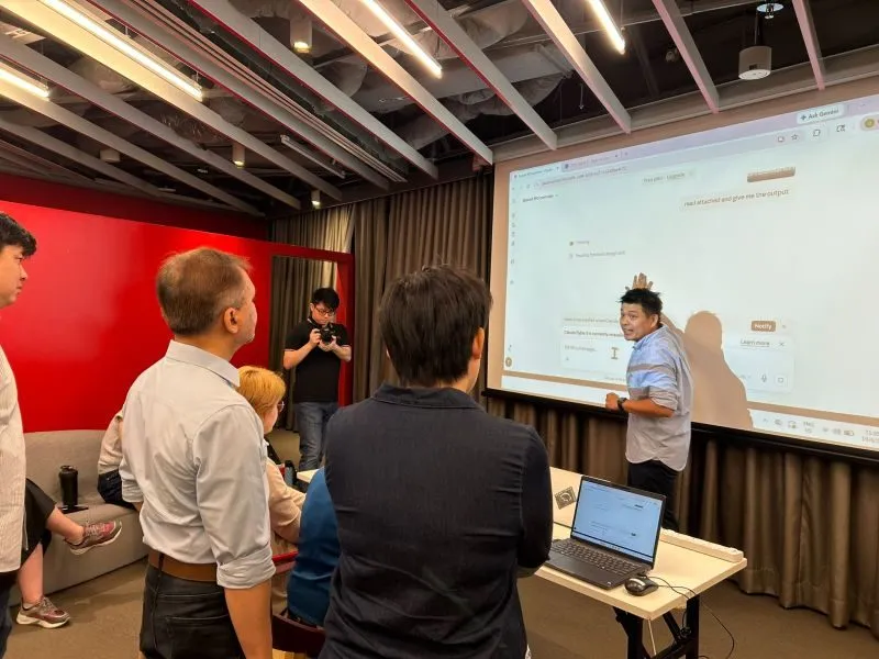 Participants gathered closely around a facilitator at a hands-on booth, watching an AI assistant work on a shared screen.