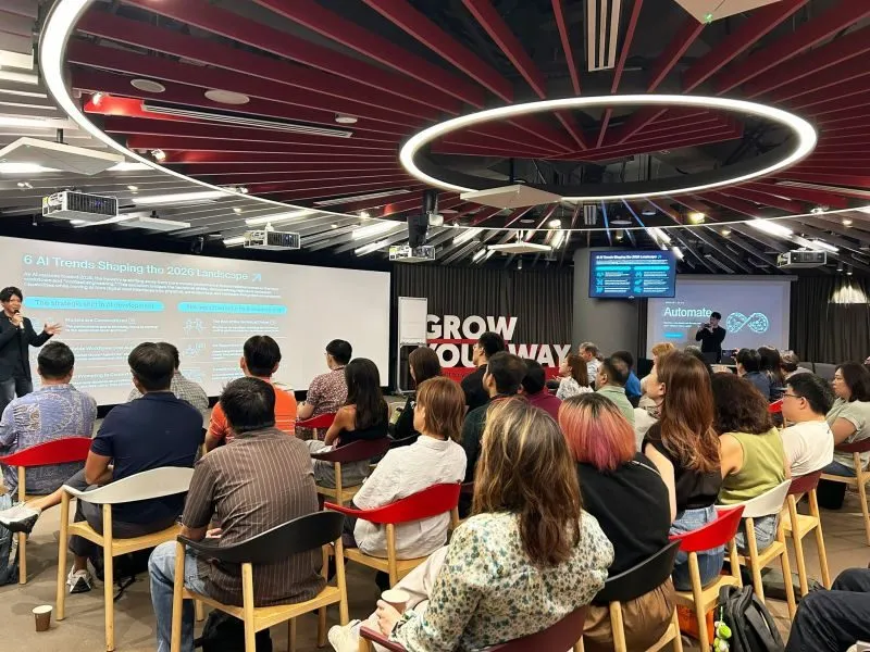 Attendees seated beneath the venue's circular ring lights, facing a presenter and a slide titled "6 AI Trends Shaping the 2026 Landscape".