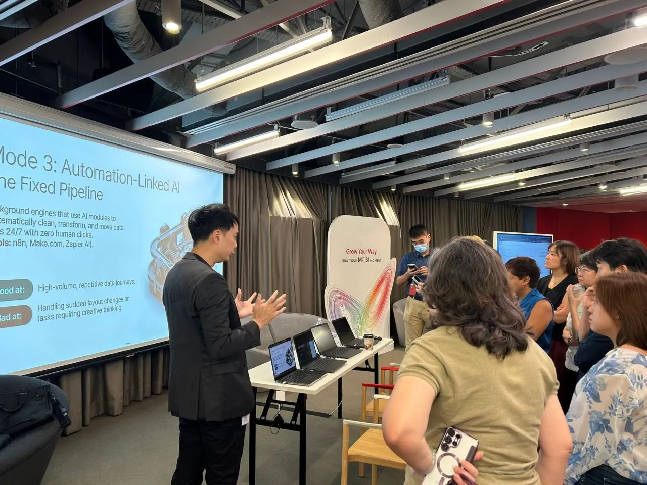 A facilitator at the automation booth explaining "Mode 3: Automation-Linked AI" with tools such as n8n, Make.com and Zapier as participants look on.