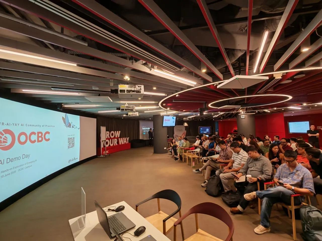 Wide view of the FR-AI-YAY AI Community of Practice Demo Day at the OCBC Campus, the event title shown on a large screen above seated attendees.